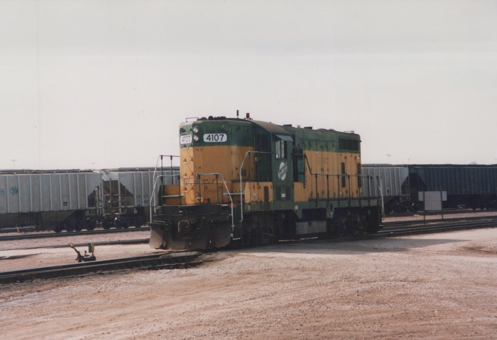 A green and yellow diesel locomotive numbered 4107 is positioned on tracks in a rail yard with freight cars in the background.
