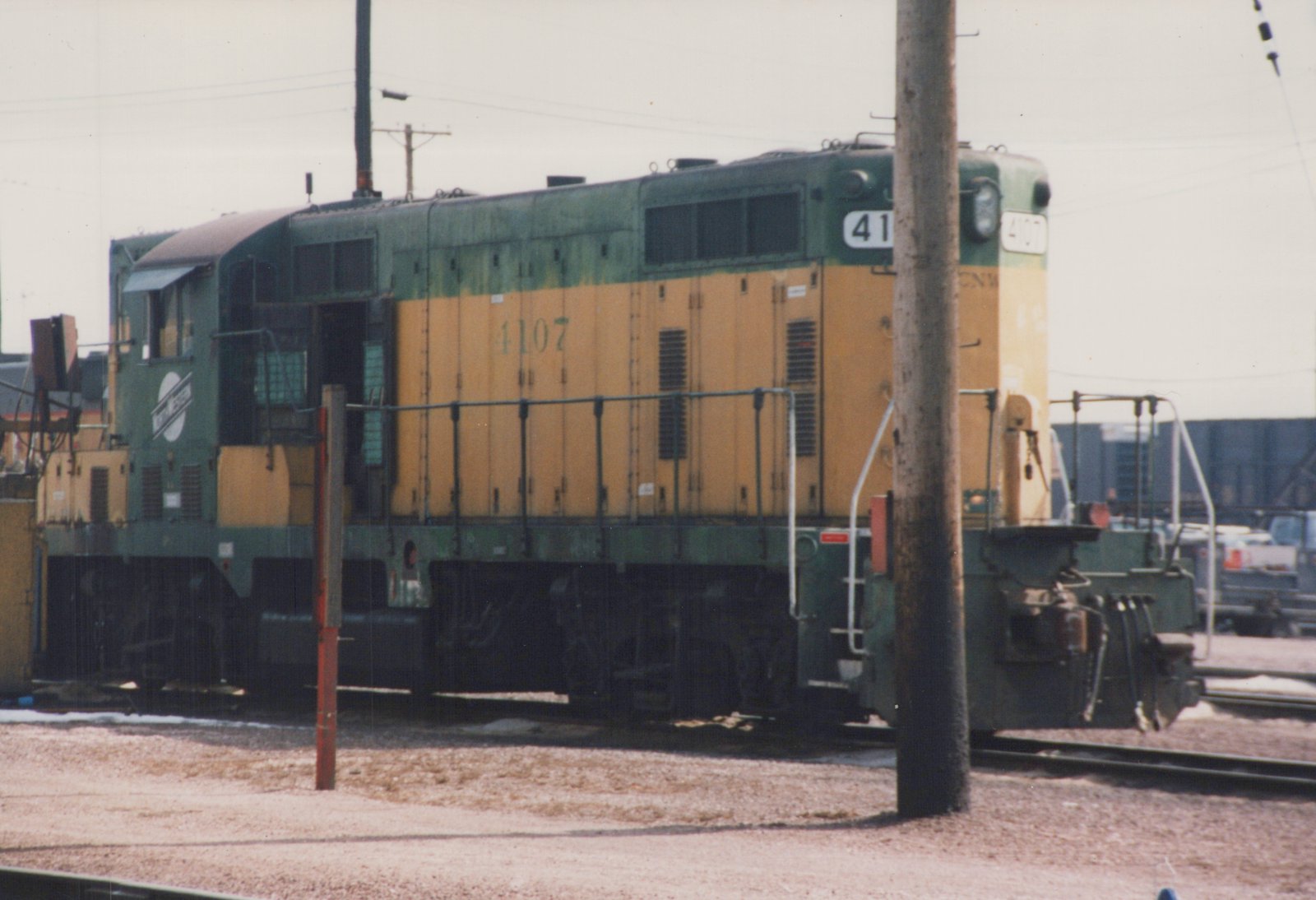 A green and yellow Chicago and North Western SW1200 locomotive, number 4107, stands on tracks in a gravel yard with snow on the ground under an overcast sky.
