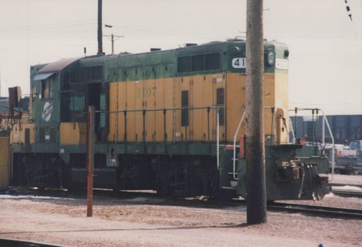 A green and yellow Chicago and North Western SW1200 locomotive, number 4107, stands on tracks in a gravel yard with snow on the ground under an overcast sky.