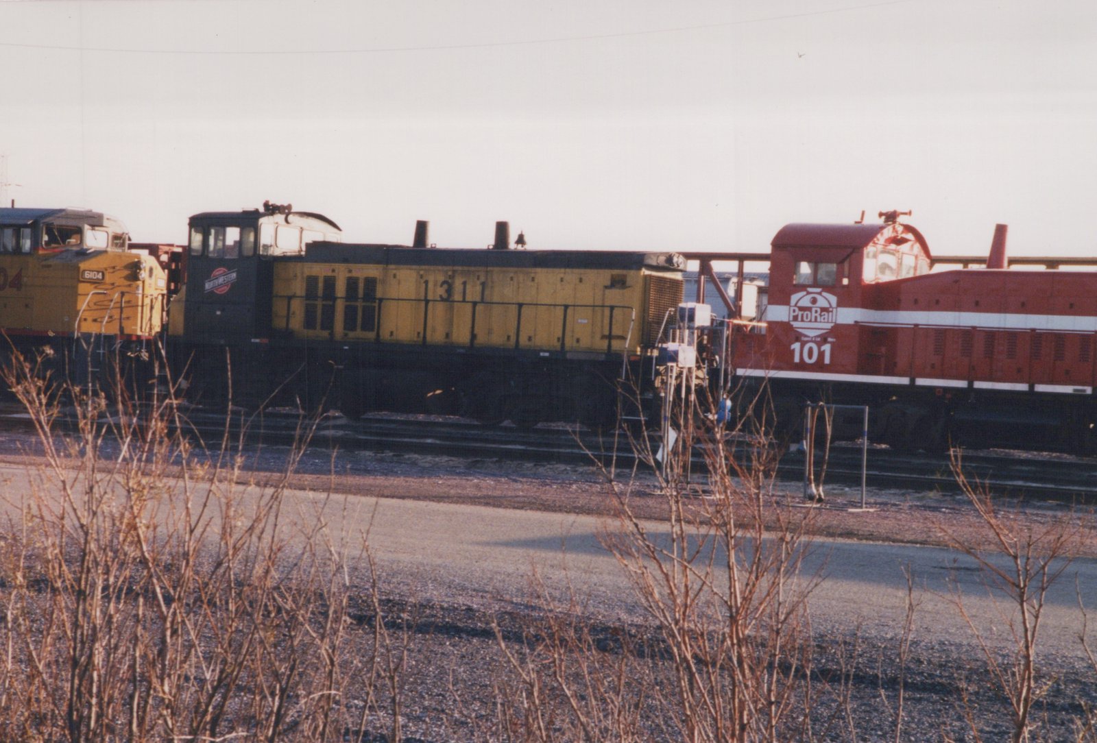 Three diesel locomotives, one yellow with black cab and number 1311, one yellow with black cab and number 6104, and one red with white stripes and number 101, are parked on tracks in a rail yard with dry brush in the foreground.