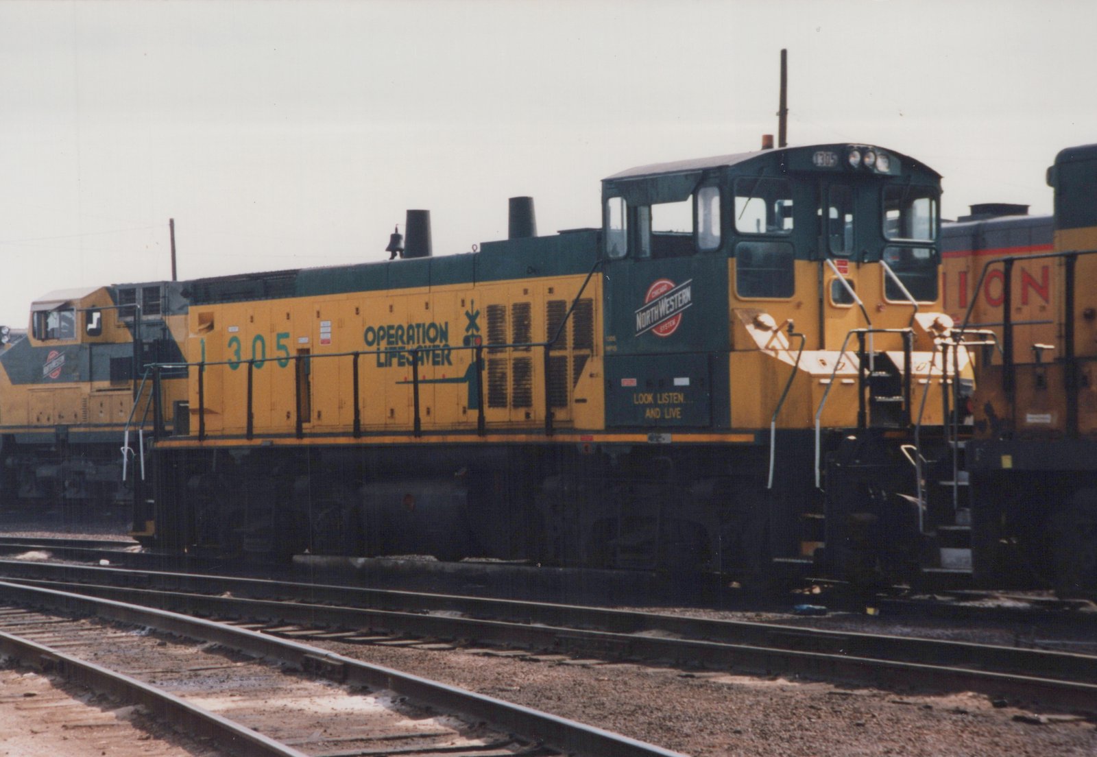 A yellow and green diesel locomotive with the number 1305 and 'Operation Lifesaver' lettering sits on railroad tracks in a yard.