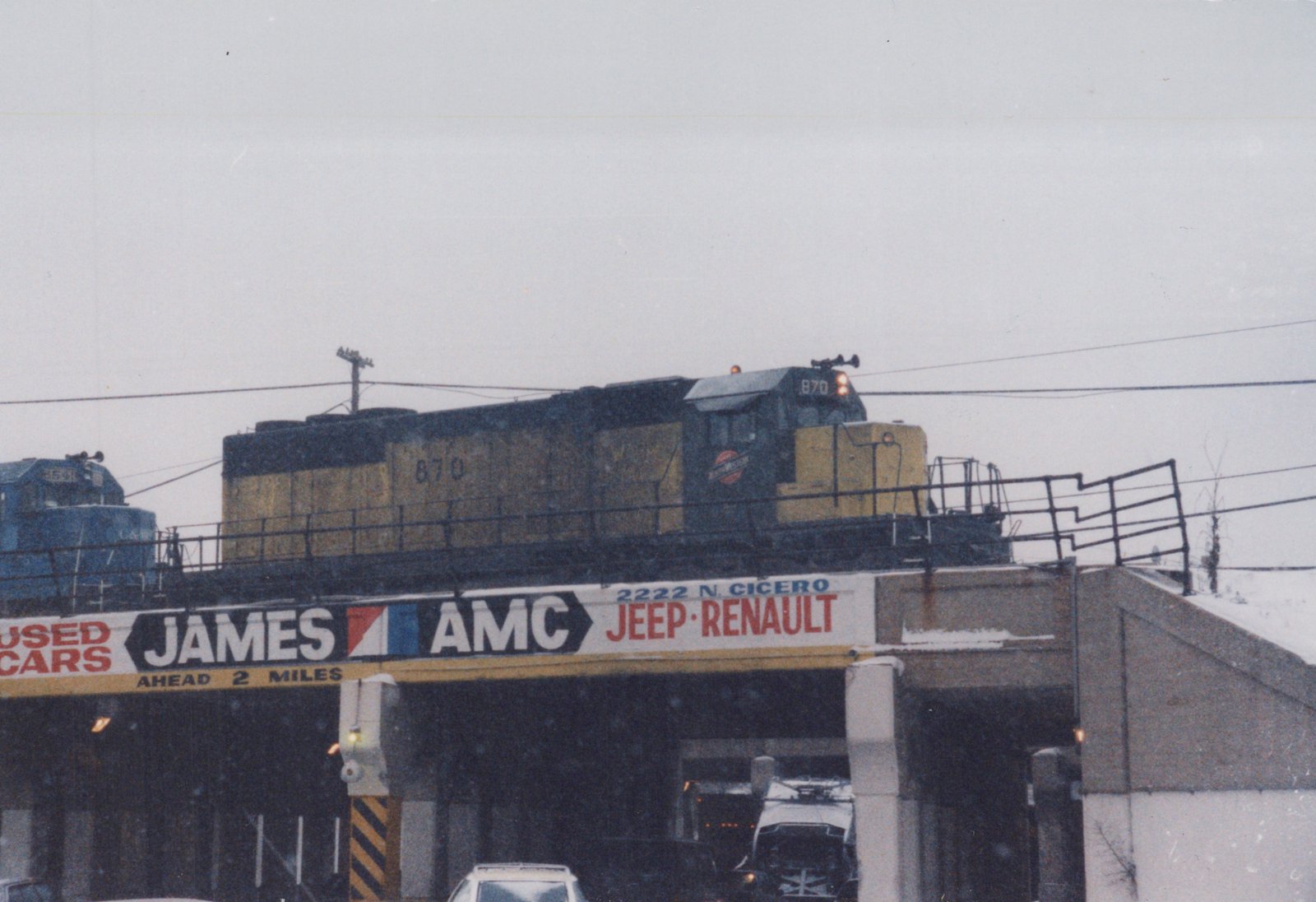 Two Chicago and North Western diesel locomotives cross an elevated overpass in snowy weather, with a trolleybus and cars visible below.