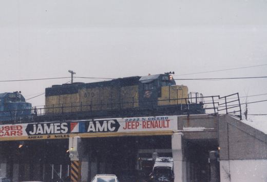 Two Chicago and North Western diesel locomotives cross an elevated overpass in snowy weather, with a trolleybus and cars visible below.