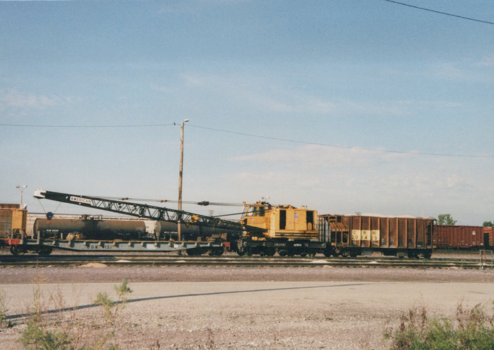 A yellow, self-propelled railway rotary crane, marked MWM 262061, is stationary in a freight yard next to an open-top Chicago and North Western gondola car with the number 201429.