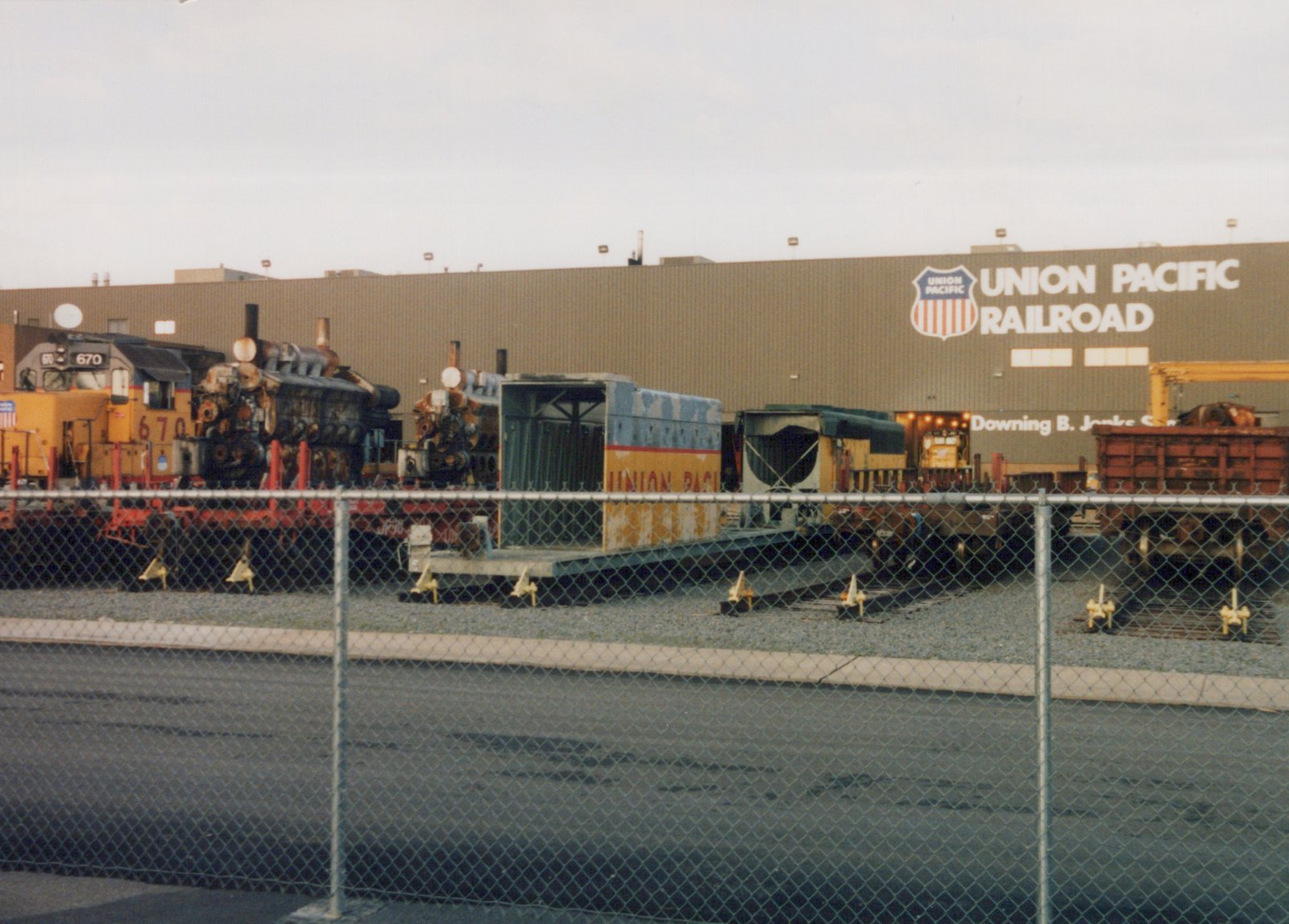 A Union Pacific diesel locomotive and parts of scrapped steam engines are displayed behind a chain-link fence in front of a large building bearing the Union Pacific Railroad name and logo.
