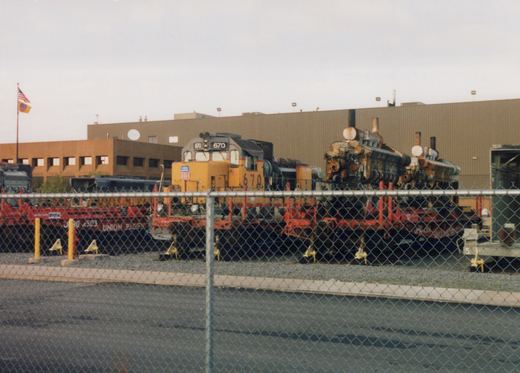 A yellow Union Pacific diesel locomotive, number 670, is seen behind a chain-link fence on a red flatcar in an industrial yard.