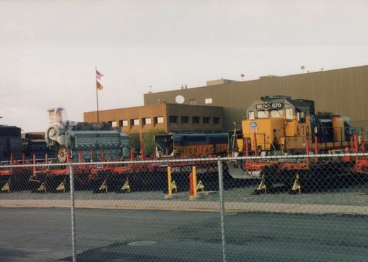 A yellow and gray Union Pacific GP9 locomotive, number 670, is parked in a rail yard with a blue SW1200 switcher and a red flatcar, with a large industrial building and an American flag in the background.
