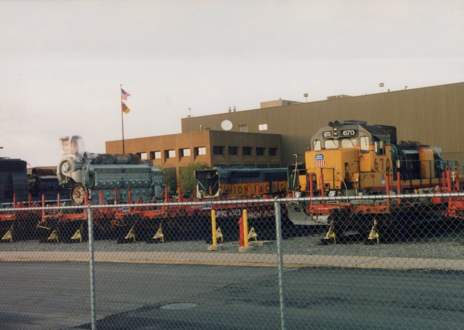 A yellow and gray Union Pacific GP9 locomotive, number 670, is parked in a rail yard with a blue SW1200 switcher and a red flatcar, with a large industrial building and an American flag in the background.