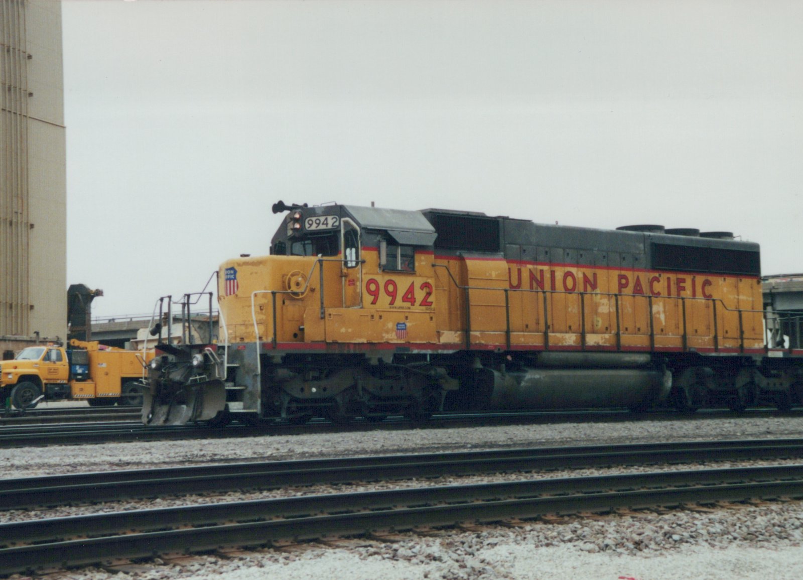 A side view of a yellow Union Pacific SD70M diesel locomotive, number 9942, on a set of tracks in a rail yard under an overcast sky.