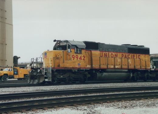 A side view of a yellow Union Pacific SD70M diesel locomotive, number 9942, on a set of tracks in a rail yard under an overcast sky.