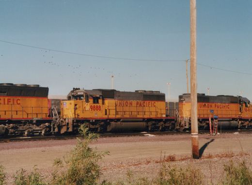 A side view of two yellow and red Union Pacific locomotives, 9888 and 9891, parked on parallel tracks in a gravel yard under a clear blue sky.