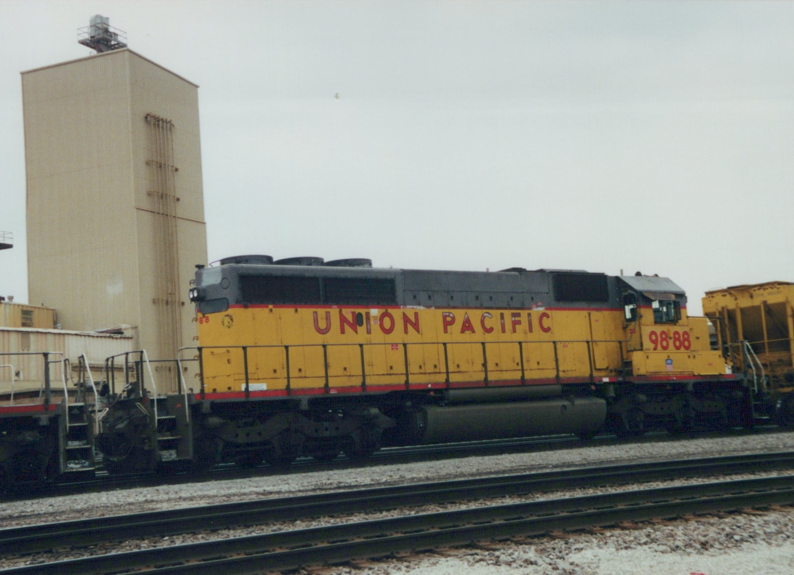 A yellow and red Union Pacific diesel locomotive, number 9888, is parked on a set of tracks in a rail yard with a tall industrial building in the background.