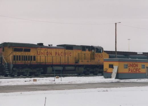 A yellow and black Union Pacific locomotive numbered 9763 stands on tracks next to a small building in a snowy yard under a gray sky.