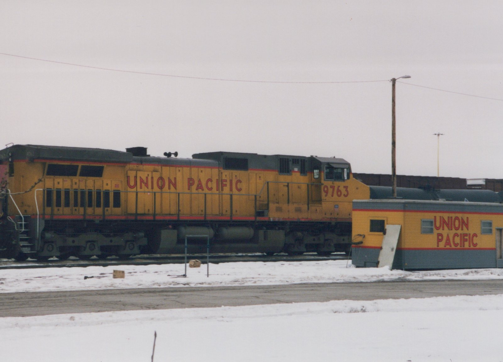 A yellow and black Union Pacific locomotive numbered 9763 stands on tracks next to a small building in a snowy yard under a gray sky.