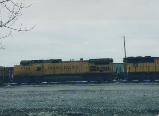 A yellow Union Pacific diesel locomotive with the number 9768 on its side sits on tracks in a gravel yard under an overcast sky, with bare tree branches visible in the top left corner.