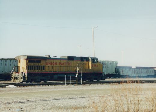 A yellow and red Union Pacific locomotive with the number 9751 is parked on railroad tracks next to blue Wisconsin Central freight cars, with dry grass in the foreground and a clear sky above.