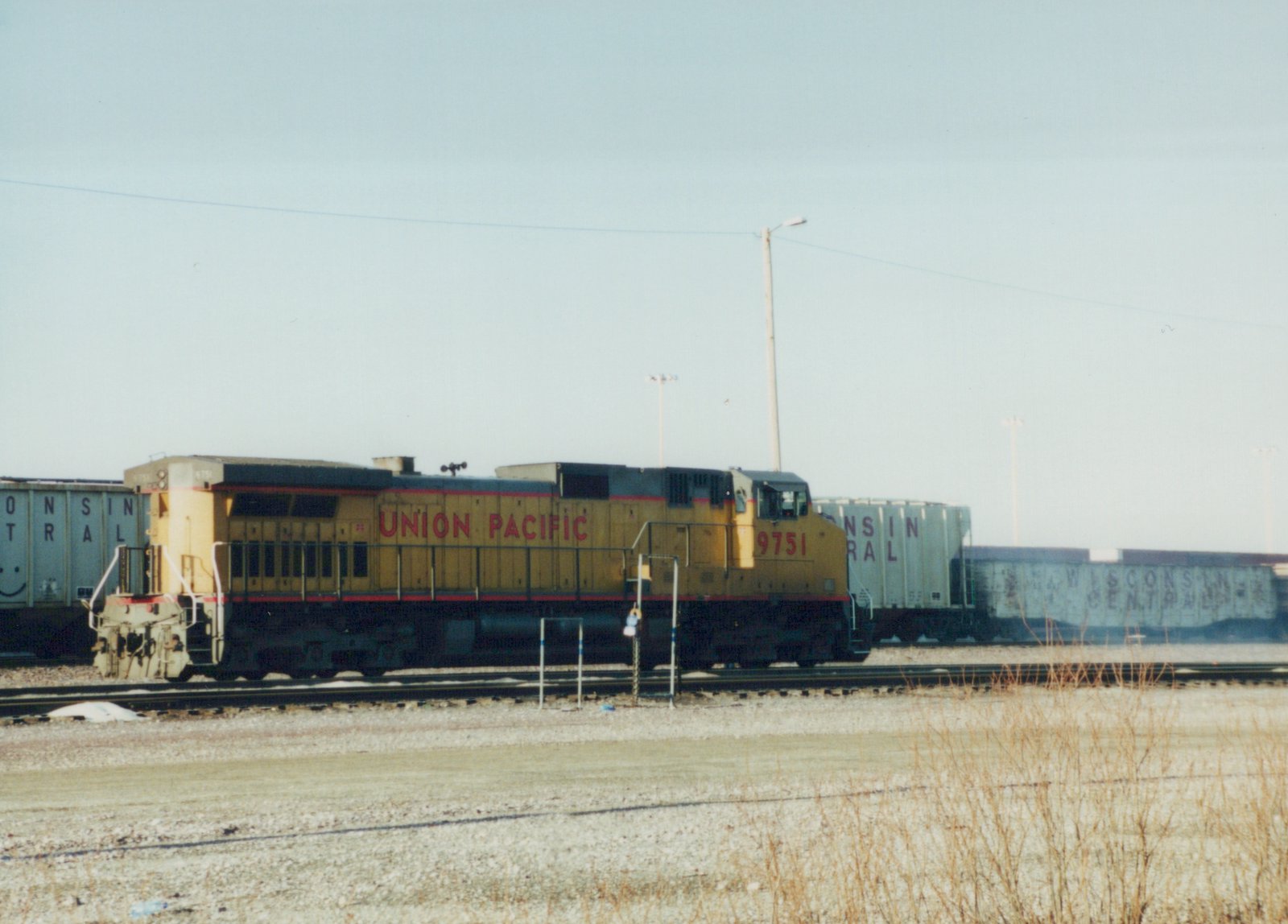 A yellow and red Union Pacific locomotive with the number 9751 is parked on railroad tracks next to blue Wisconsin Central freight cars, with dry grass in the foreground and a clear sky above.