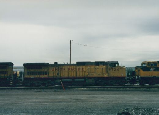 A side view of a yellow Union Pacific diesel locomotive, number 9734, parked in a gravel rail yard with other locomotives nearby under a cloudy sky.
