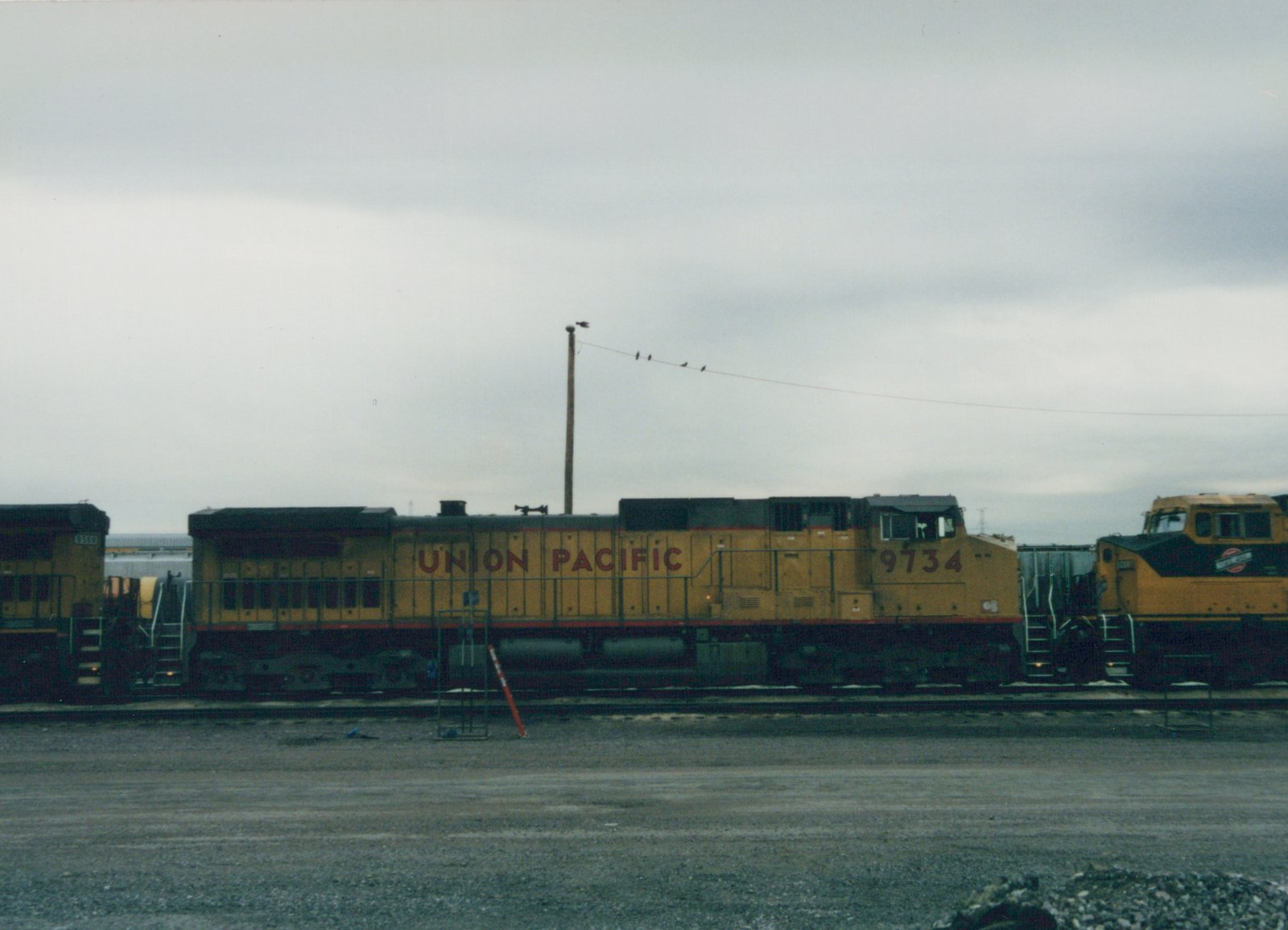 A side view of a yellow Union Pacific diesel locomotive, number 9734, parked in a gravel rail yard with other locomotives nearby under a cloudy sky.