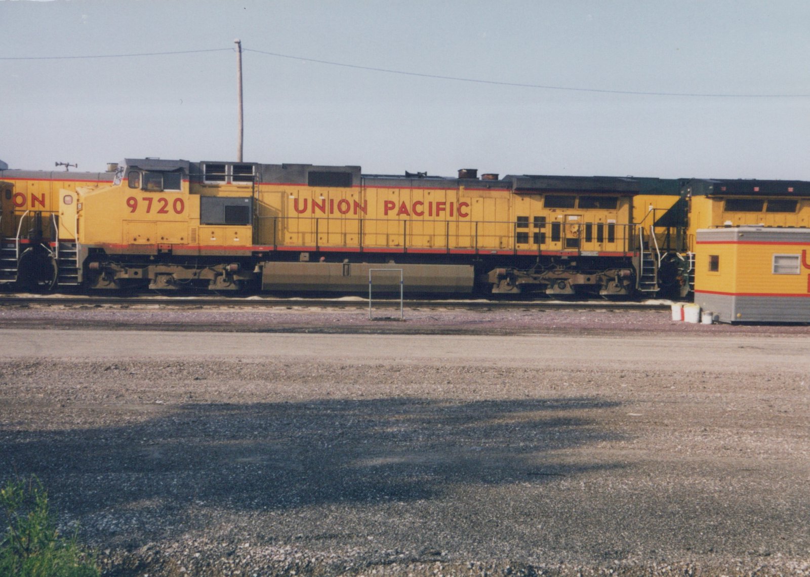 A side view of a yellow Union Pacific diesel locomotive, number 9720, parked on tracks beside a gravel lot, with another locomotive and a yellow and gray trailer behind it.