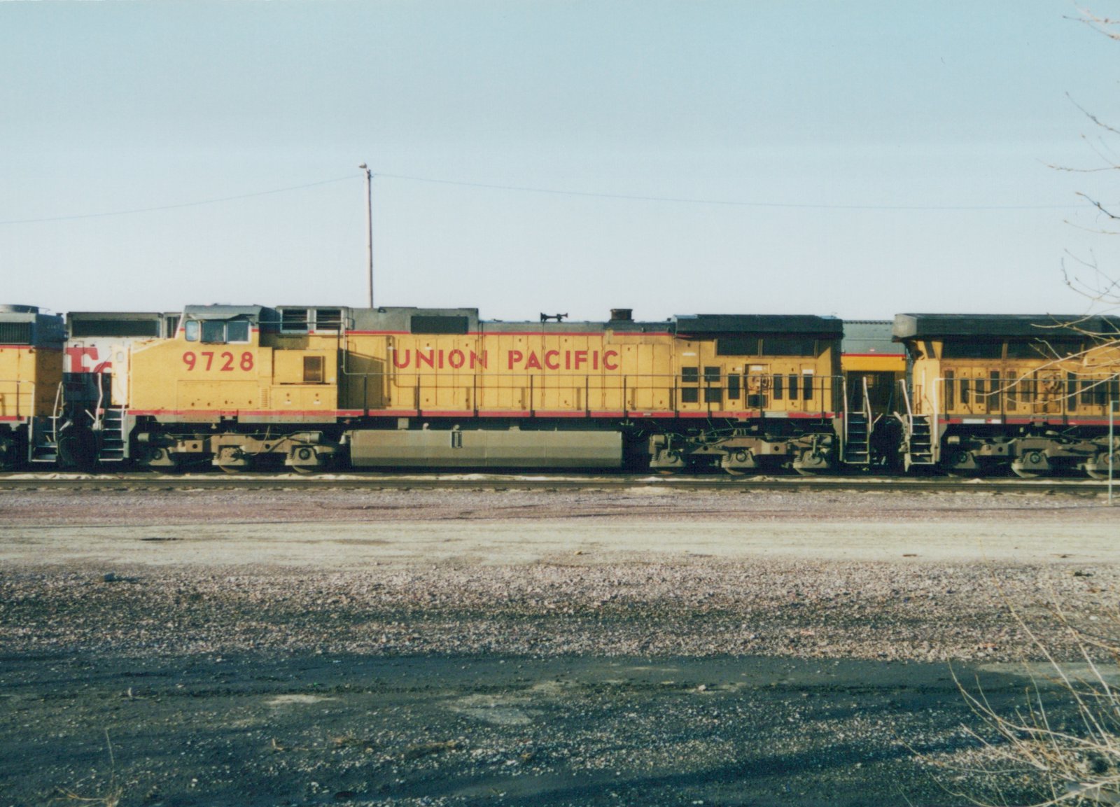 Side view of a yellow and red Union Pacific diesel locomotive numbered 9728, with two other locomotives attached, sitting on tracks in a gravel yard under a clear sky.