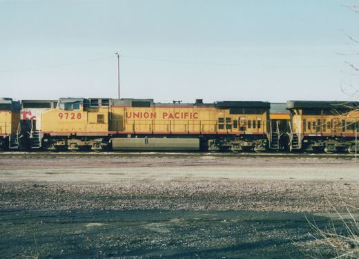 Side view of a yellow and red Union Pacific diesel locomotive numbered 9728, with two other locomotives attached, sitting on tracks in a gravel yard under a clear sky.