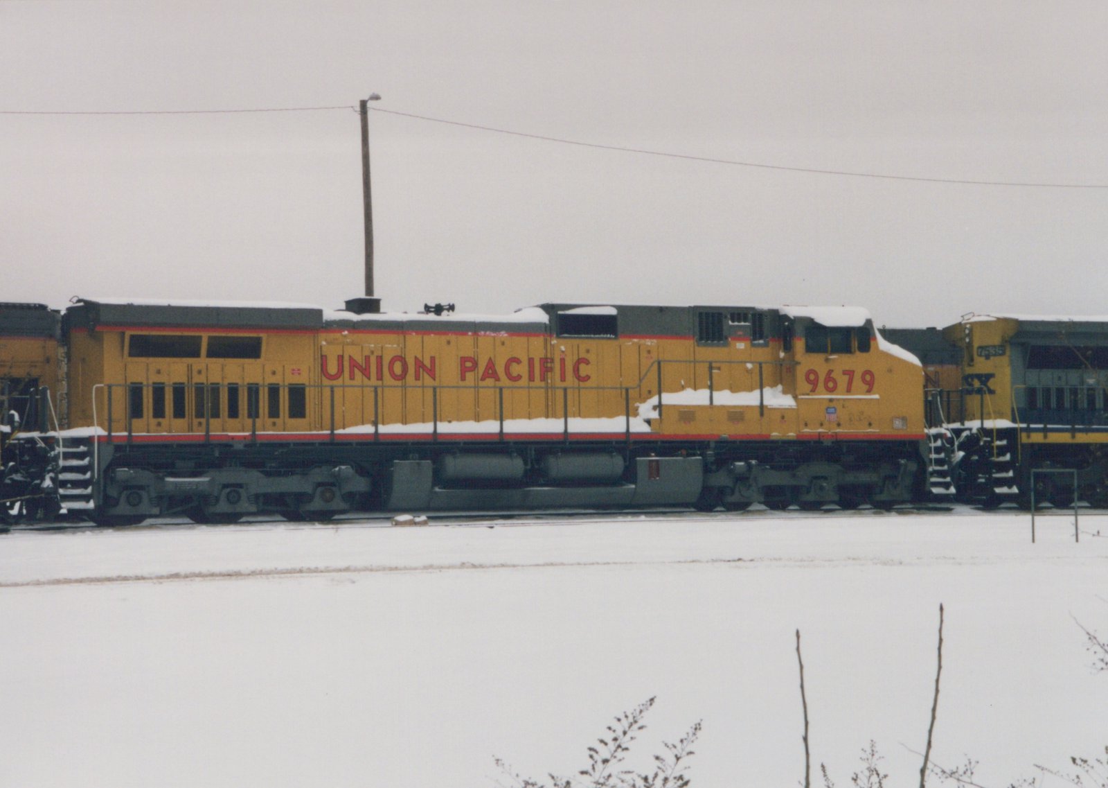 A side view of a yellow Union Pacific diesel locomotive numbered 9679, standing on tracks in a snowy yard next to another diesel locomotive with the reporting mark SX.