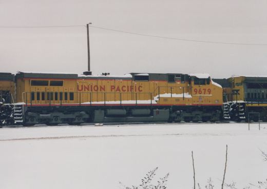 A side view of a yellow Union Pacific diesel locomotive numbered 9679, standing on tracks in a snowy yard next to another diesel locomotive with the reporting mark SX.