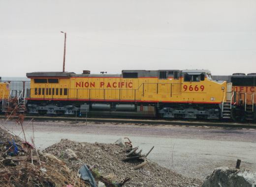 A yellow Union Pacific diesel locomotive numbered 9669 is parked on tracks in a railroad yard, with overcast skies and some debris in the foreground.