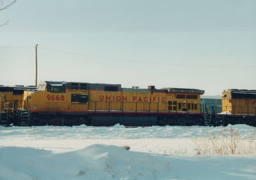 A side view of a yellow and red Union Pacific locomotive numbered 9668 in a snow-covered railroad yard, with other trains visible in the background.