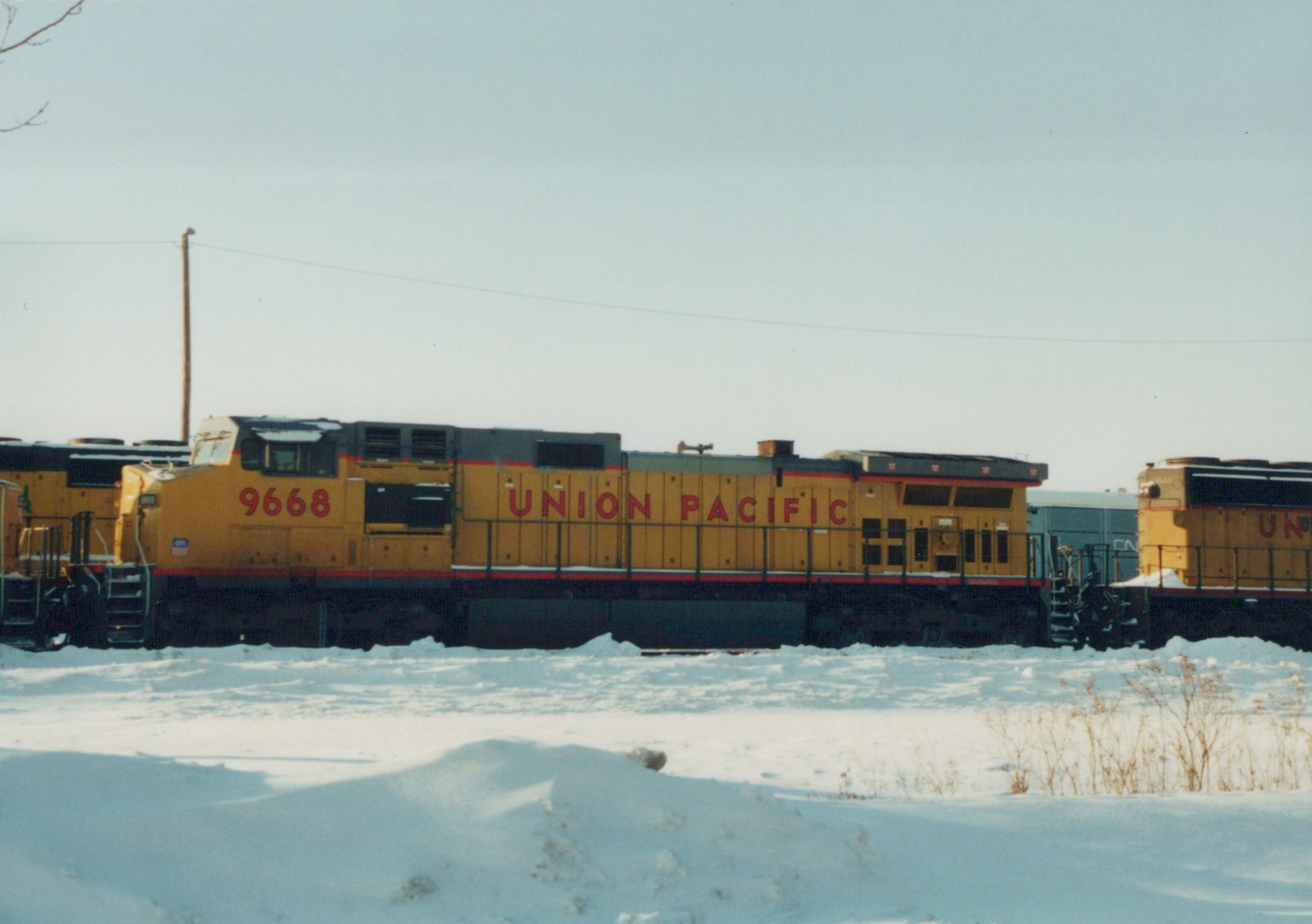 A side view of a yellow and red Union Pacific locomotive numbered 9668 in a snow-covered railroad yard, with other trains visible in the background.