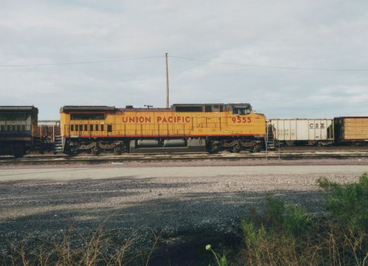 A yellow Union Pacific diesel locomotive with the number 9555 is positioned on railroad tracks next to white CSX freight cars in a gravel yard with an overcast sky and some greenery in the foreground.