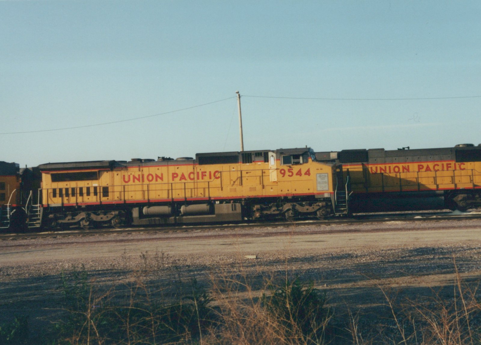 Side view of a yellow and red Union Pacific diesel locomotive numbered 9544 on a railroad track with another locomotive coupled behind it, set against a clear sky with dry grass in the foreground.