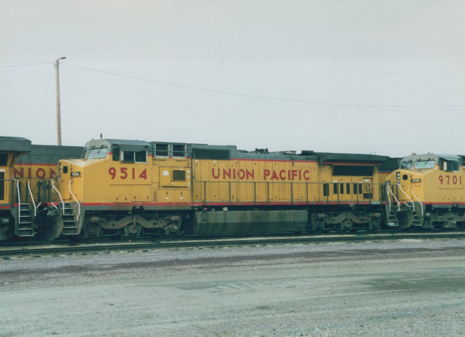 Side view of two yellow Union Pacific diesel locomotives, numbered 9514 and 9701, standing on tracks in a gravel yard under a gray sky.