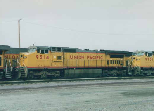 Side view of two yellow Union Pacific diesel locomotives, numbered 9514 and 9701, standing on tracks in a gravel yard under a gray sky.