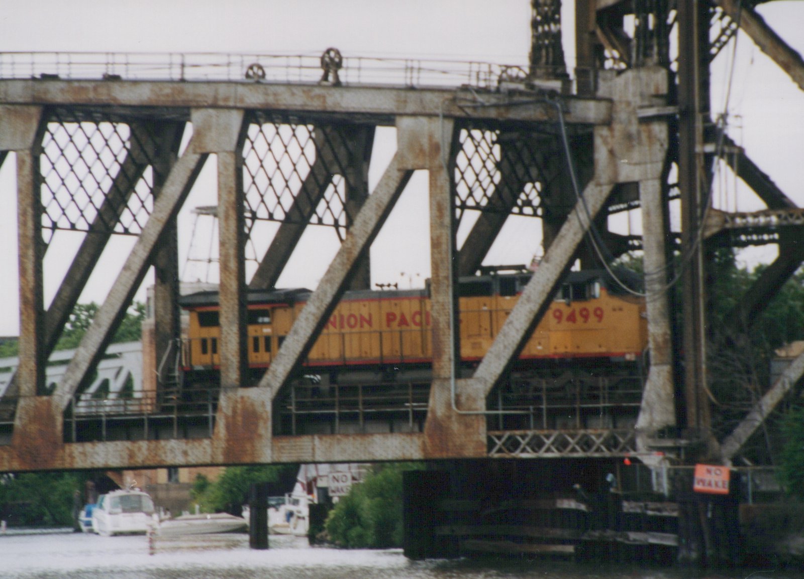 A yellow Union Pacific diesel locomotive, number 9499, is seen crossing a metal truss bridge over a river with boats below.