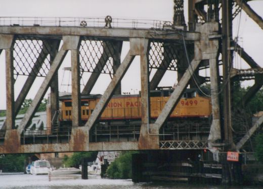 A yellow Union Pacific diesel locomotive, number 9499, is seen crossing a metal truss bridge over a river with boats below.
