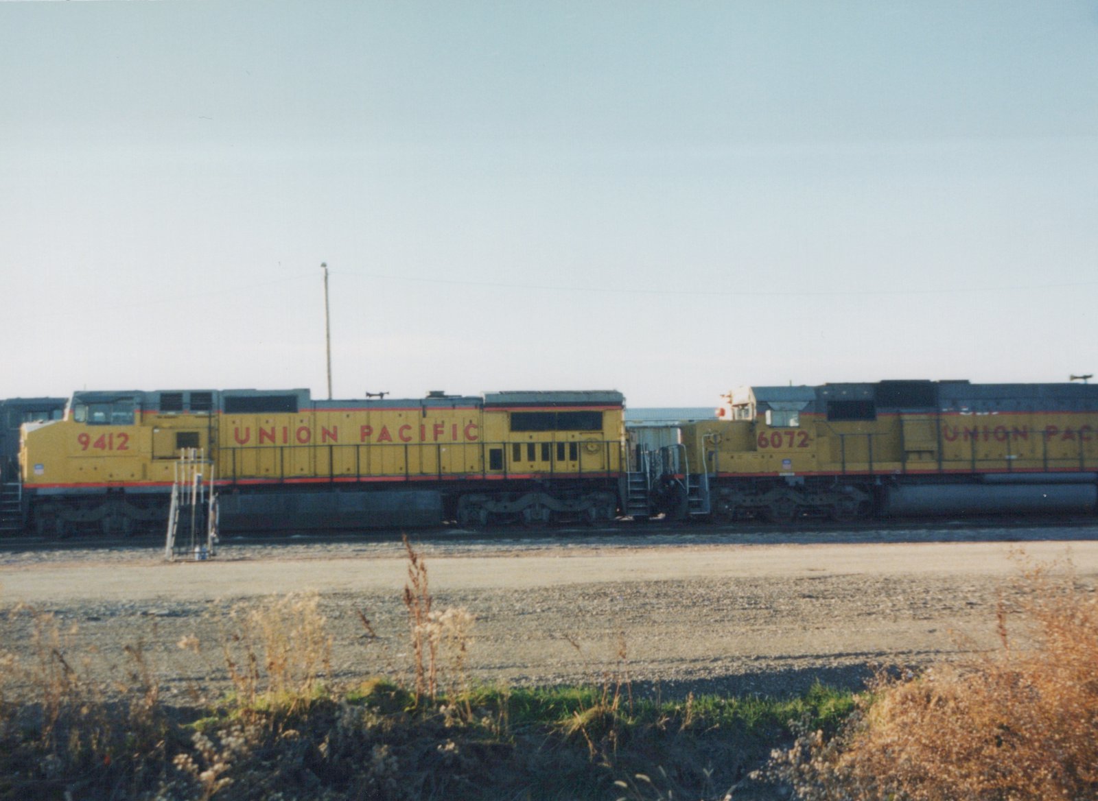 Two yellow Union Pacific diesel locomotives, numbers 9412 and 6072, sit side-by-side on tracks in a gravel yard with dry grass in the foreground on a clear autumn day.
