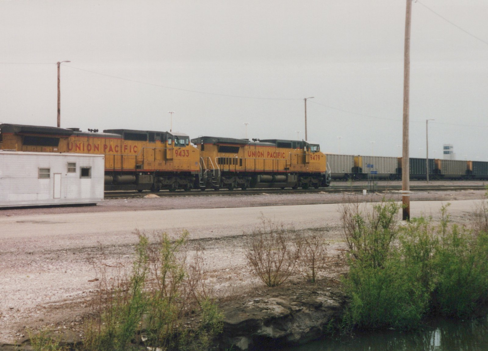 Two yellow Union Pacific locomotives, numbers 9433 and 9479, lead a freight train on tracks next to a gravel lot and a small pond under an overcast sky.