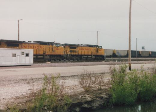 Two yellow Union Pacific locomotives, numbers 9433 and 9479, lead a freight train on tracks next to a gravel lot and a small pond under an overcast sky.
