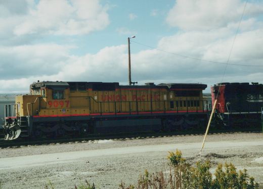 A yellow and black Union Pacific diesel locomotive, number 9097, is parked on tracks in a gravel yard with another locomotive partially visible behind it.