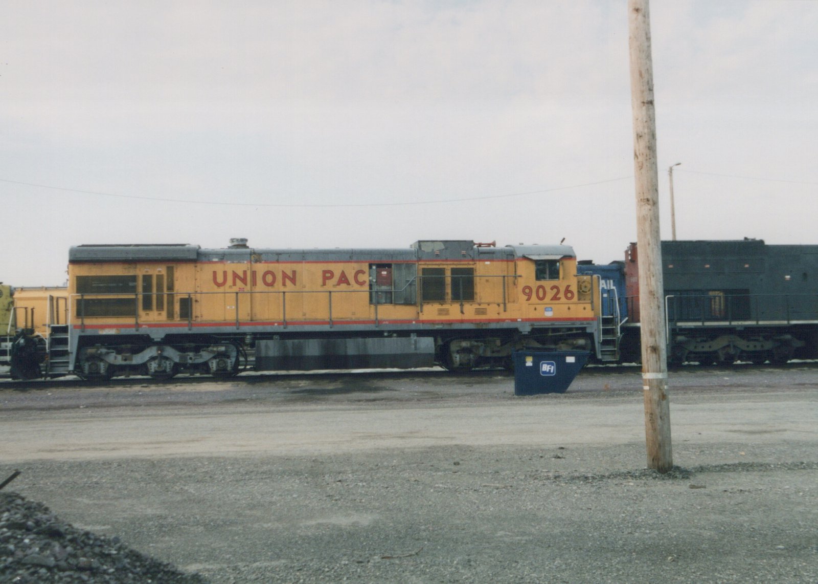 A yellow and gray Union Pacific diesel locomotive, number 9026, is parked on a railroad track in a gravel yard, with a blue dumpster and a wooden pole in the foreground.