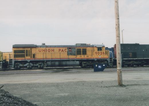 A yellow and gray Union Pacific diesel locomotive, number 9026, is parked on a railroad track in a gravel yard, with a blue dumpster and a wooden pole in the foreground.