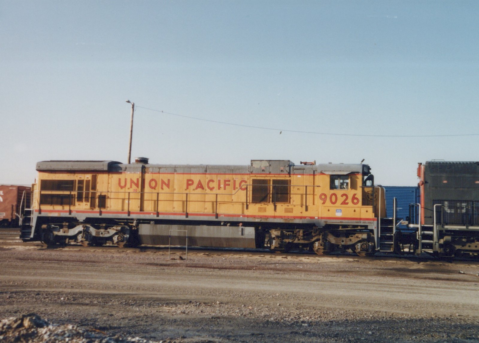 A yellow Union Pacific locomotive numbered 9026 stands in a rail yard next to a gray and blue freight car under a clear sky.