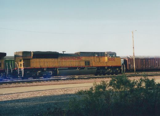 A Union Pacific diesel locomotive with the number 8550 is parked on a set of railroad tracks next to a brown freight car, with bushes in the foreground and a clear sky above.