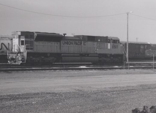 A black-and-white photo of a Union Pacific locomotive, number 8507, standing on tracks in a gravel yard with snow on the ground.