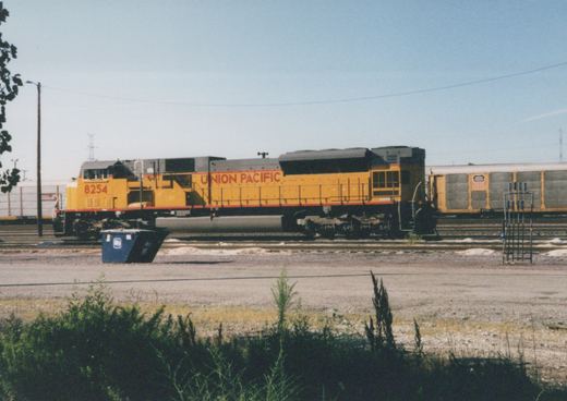 A yellow Union Pacific diesel locomotive numbered 8254 is parked in a rail yard next to a freight car, with a clear blue sky overhead and some green brush in the foreground.
