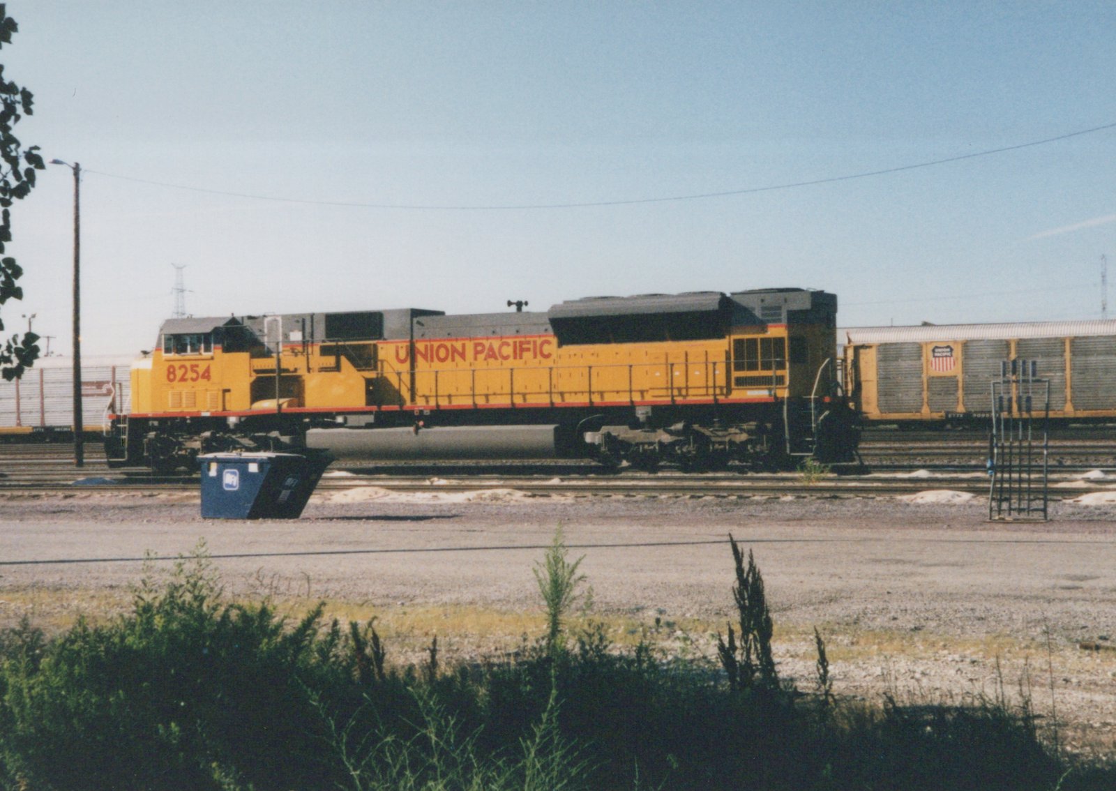 A yellow Union Pacific diesel locomotive numbered 8254 is parked in a rail yard next to a freight car, with a clear blue sky overhead and some green brush in the foreground.