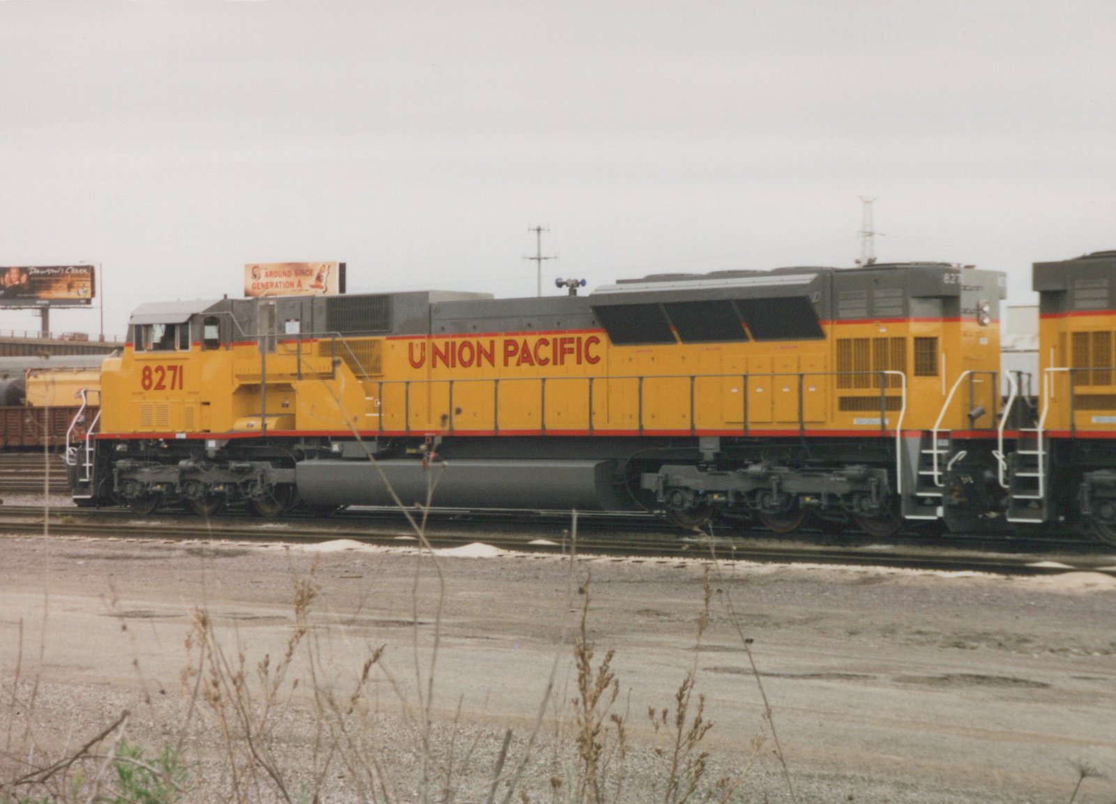 A yellow and gray Union Pacific locomotive numbered 8271 is parked on a track in a gravel yard under an overcast sky, with billboards visible in the background.