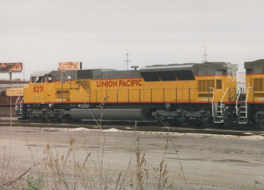 A yellow and gray Union Pacific locomotive numbered 8271 is parked on a track in a gravel yard under an overcast sky, with billboards visible in the background.